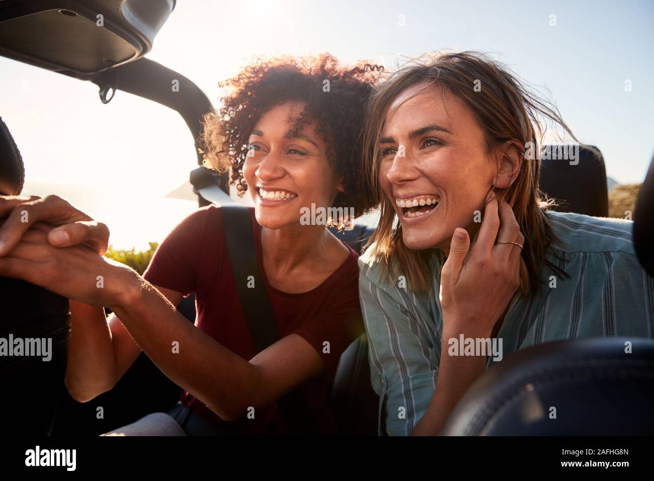 Two millennial female friends on a road trip smiling in the back of an open top car, close up Stock Photo