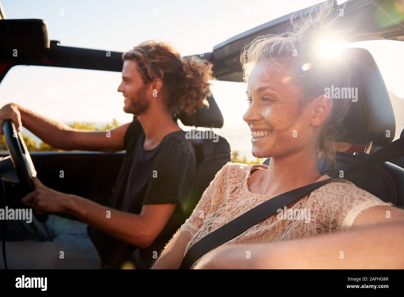 Millennial white couple on a road trip driving in an open top car, backlit, close up Stock Photo