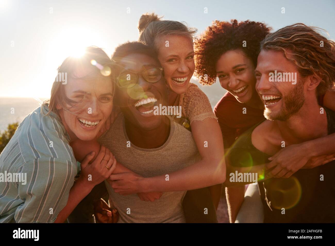 Five millennial friends on a road trip have fun piggybacking at the roadside, front view, lens flare Stock Photo