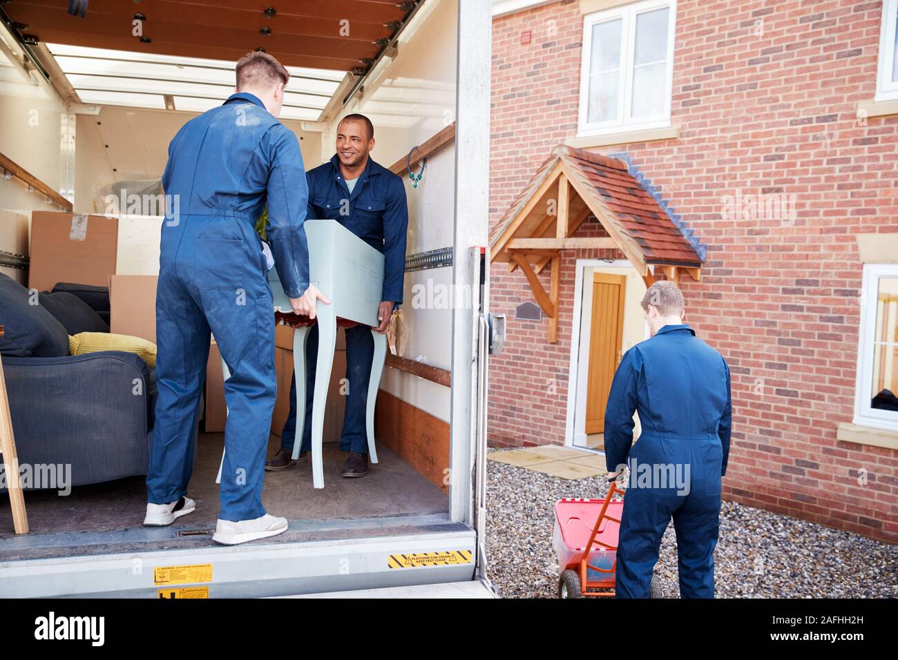 Removal Company Workers Unloading Furniture And Boxes From Truck Into New Home On Moving Day Stock Photo