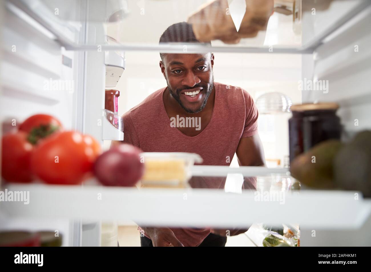 View Looking Out From Inside Of Refrigerator As Man Opens Door And Unpacks Shopping Bag Of Food Stock Photo