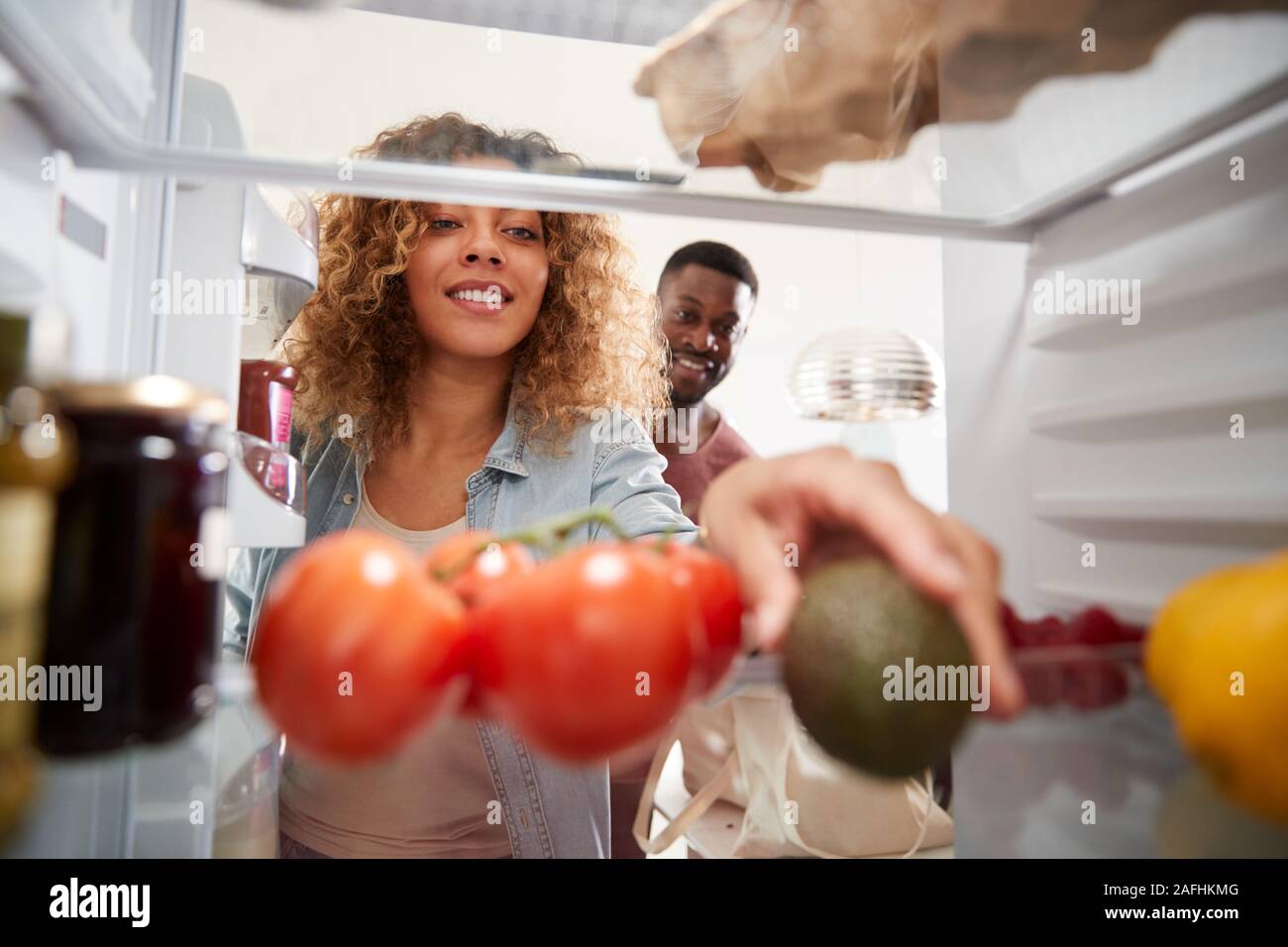 View Looking Out From Inside Of Refrigerator As Couple Open Door And Unpack Shopping Bag Of Food Stock Photo