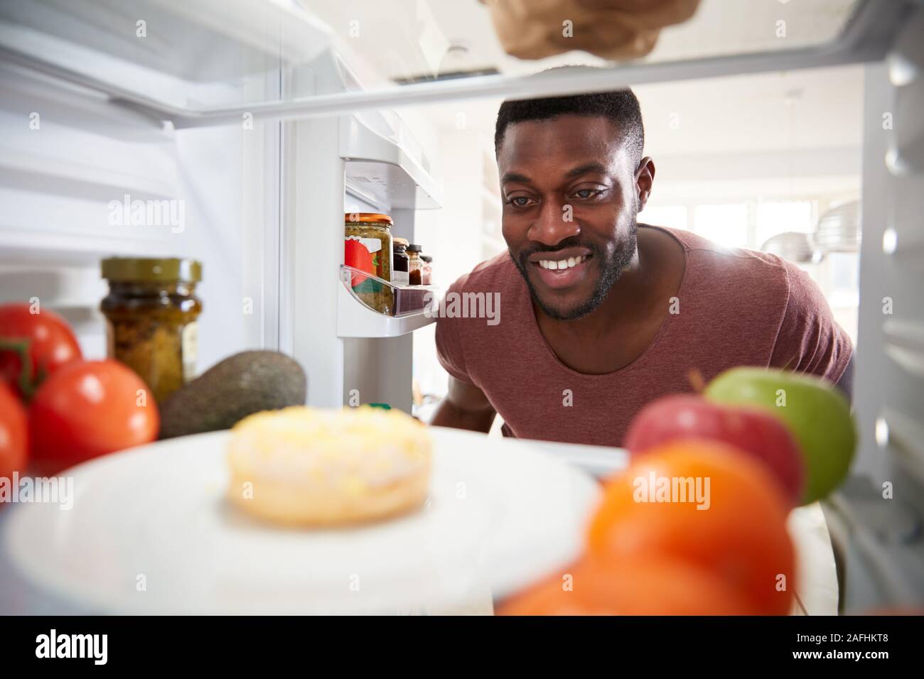 View Looking Out From Inside Of Refrigerator As Man Opens Door And Reaches For Unhealthy Donut Stock Photo