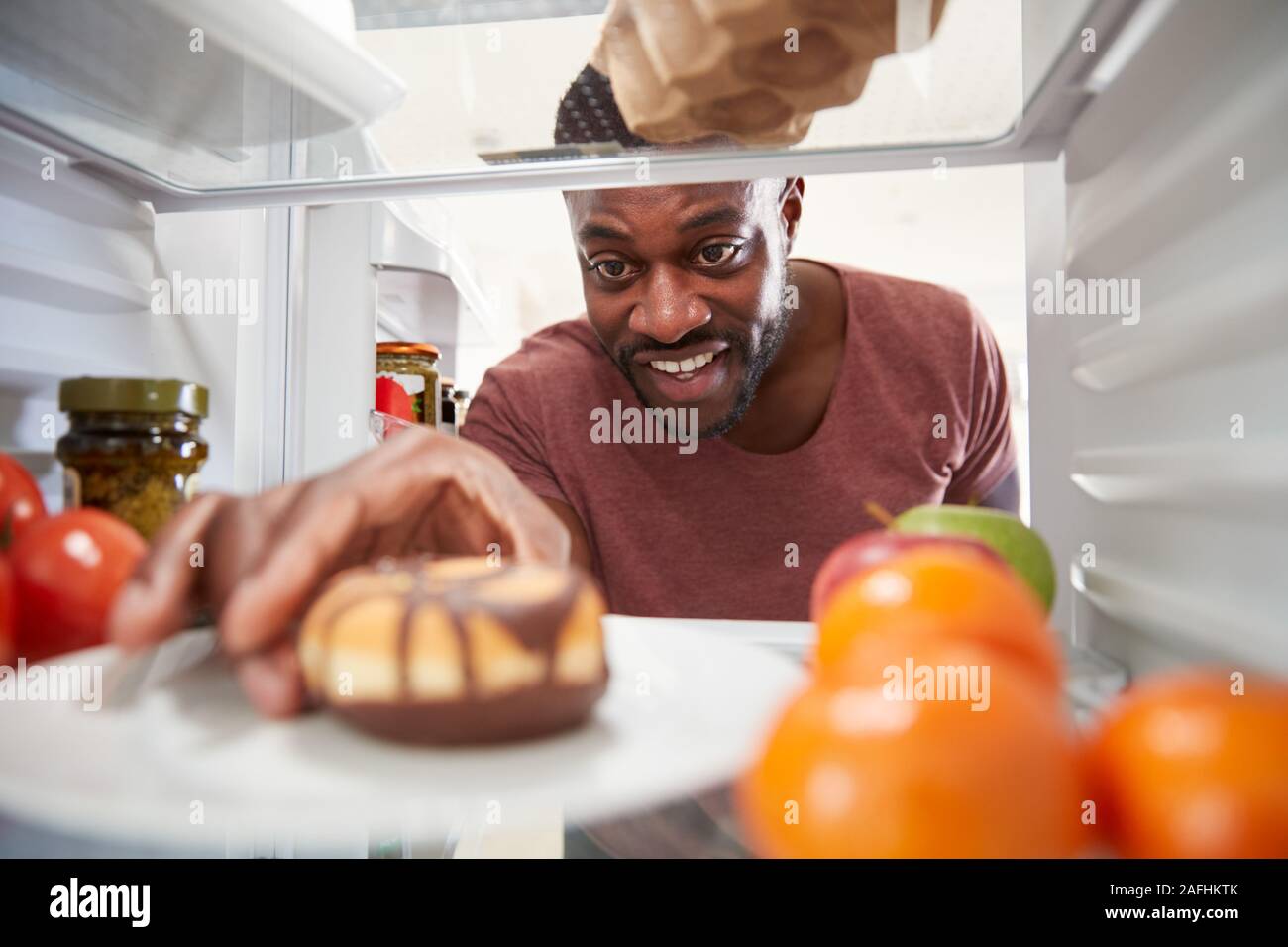 View Looking Out From Inside Of Refrigerator As Man Opens Door And Reaches For Unhealthy Donut Stock Photo