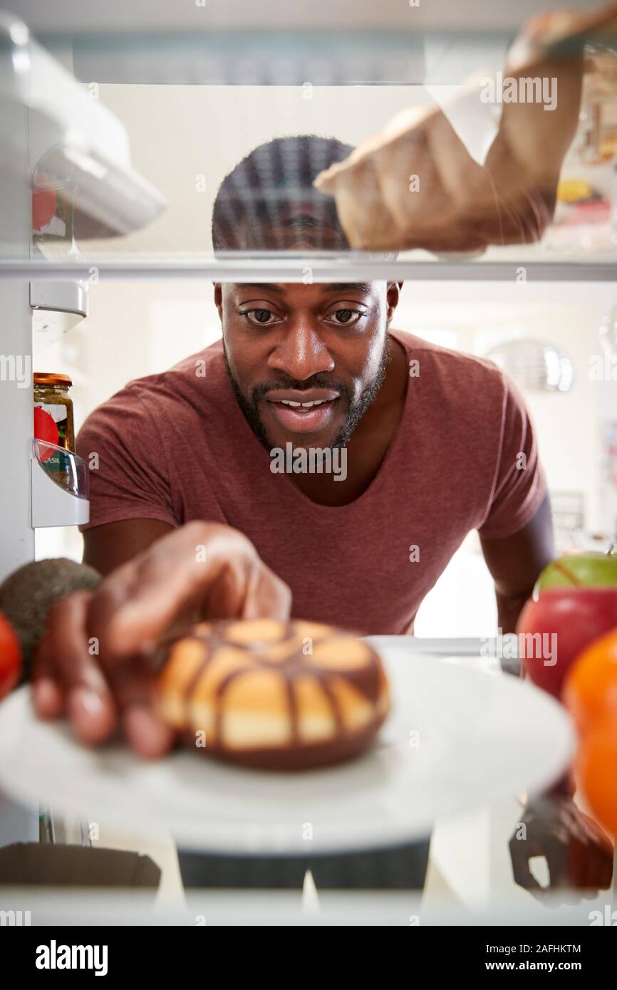 View Looking Out From Inside Of Refrigerator As Man Opens Door And Reaches For Unhealthy Donut Stock Photo