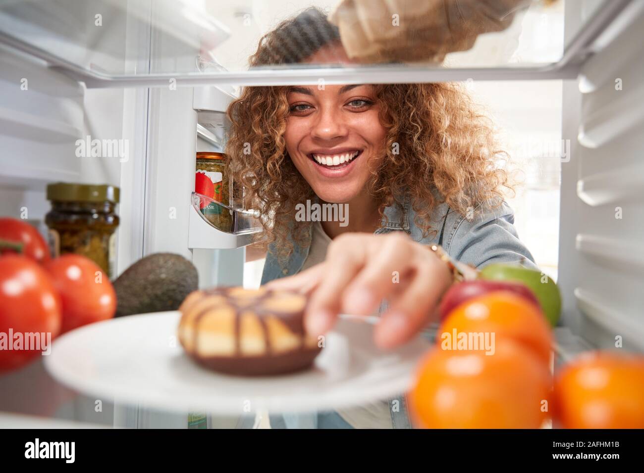 View Looking Out From Inside Of Refrigerator As Woman Opens Door And Reaches For Unhealthy Donut Stock Photo