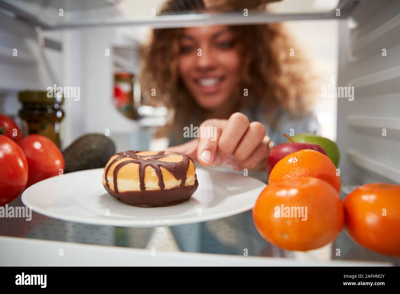 View Looking Out From Inside Of Refrigerator As Woman Opens Door And Reaches For Unhealthy Donut Stock Photo