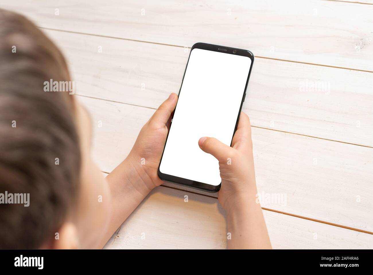 Kid holding smart phone and touch isolated display for app, or game promotion. White wooden desk in background. Modern smart phone with round edges Stock Photo