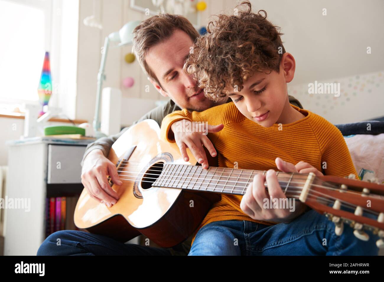 Single Father At Home With Son Teaching Him To Play Acoustic Guitar In Bedroom Stock Photo