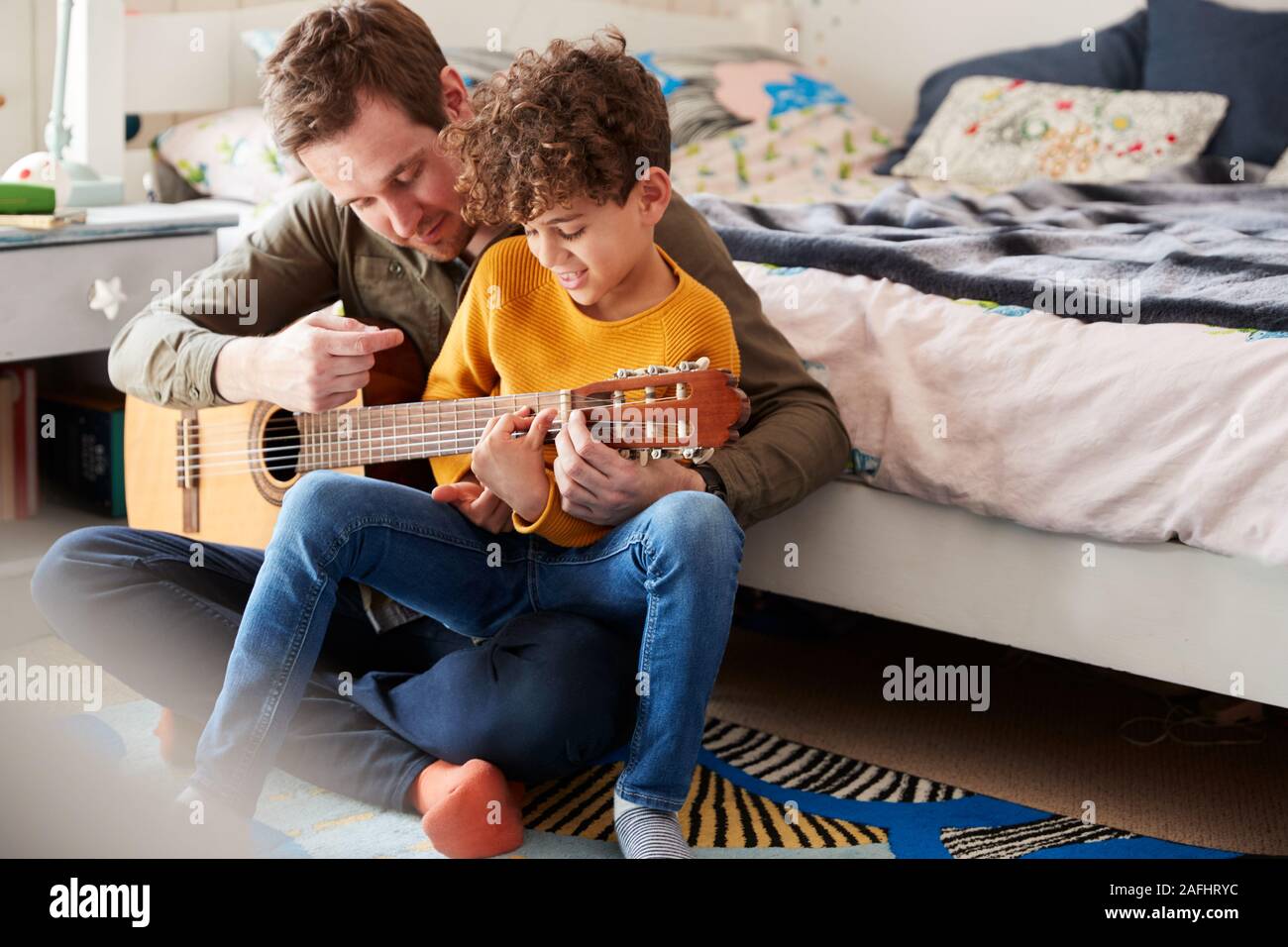Single Father At Home With Son Teaching Him To Play Acoustic Guitar In Bedroom Stock Photo