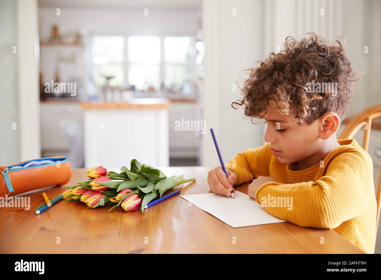 Young Boy At Home With Bunch Of Flowers Writing In Mothers Day Card Stock Photo