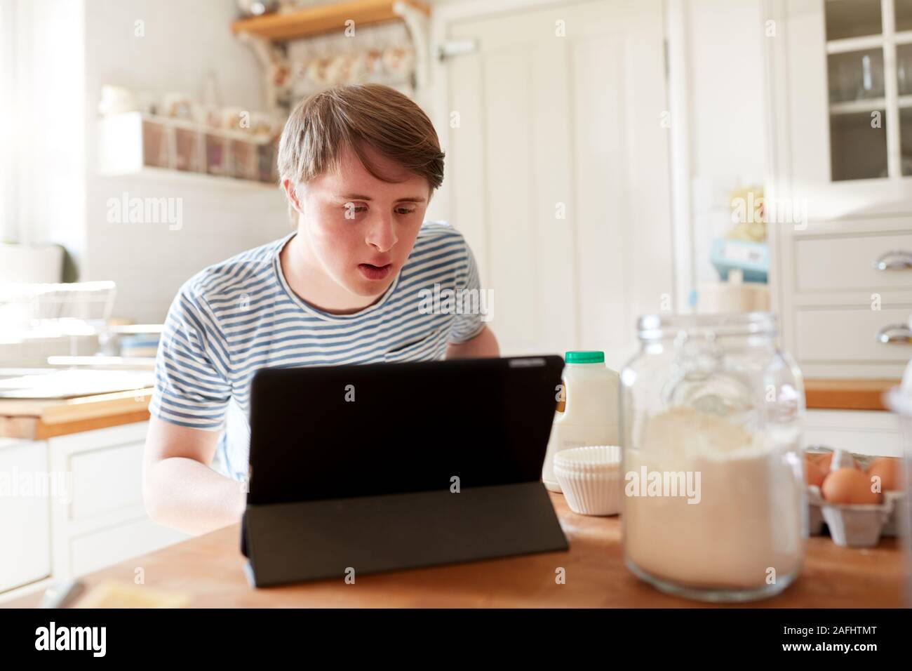 Young Man With Downs Syndrome Following Recipe On Digital Tablet To Bake Cake In Kitchen At Home Stock Photo