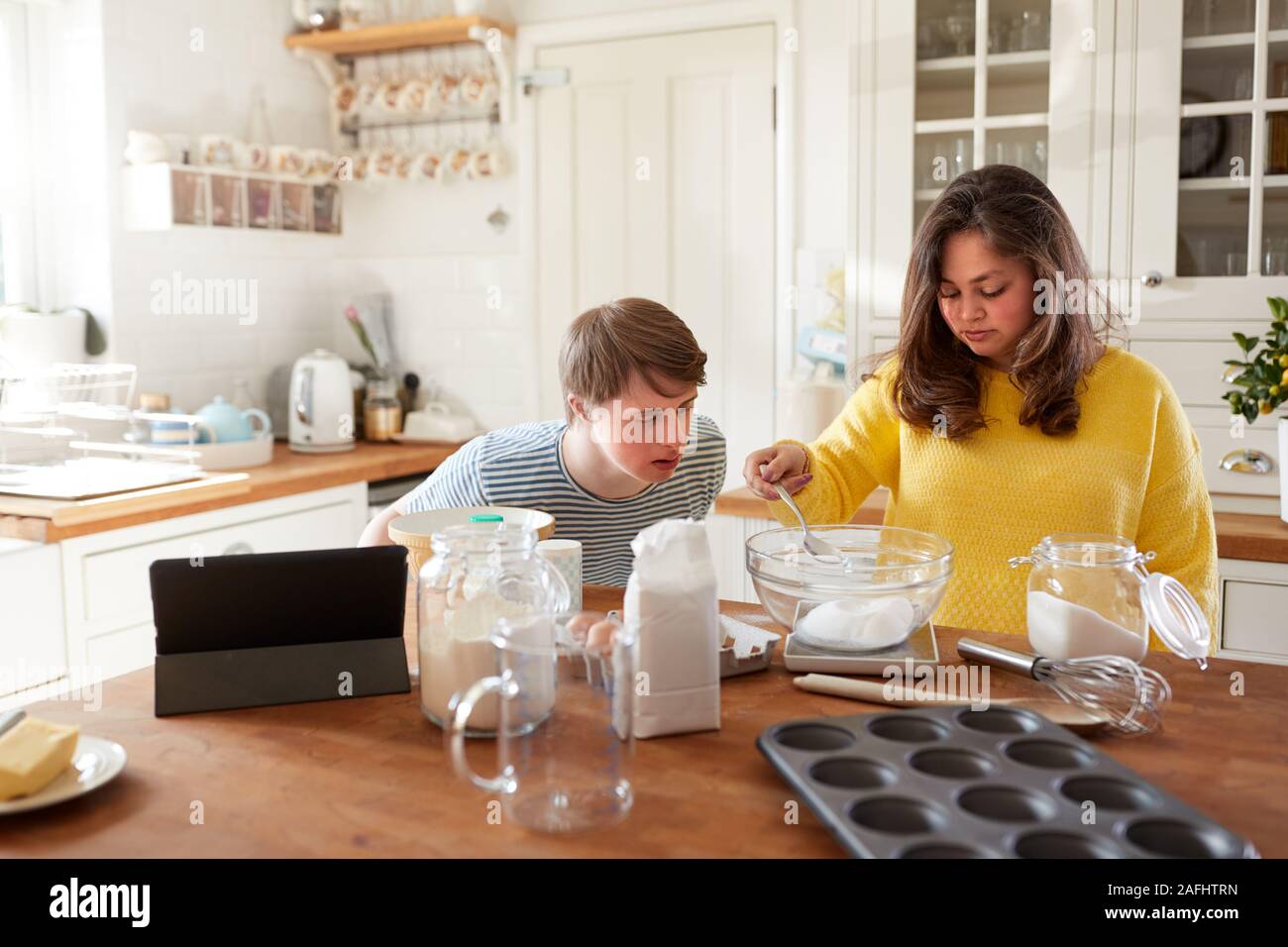 Young Downs Syndrome Couple Following Recipe On Digital Tablet To Bake Cake In Kitchen At Home Stock Photo