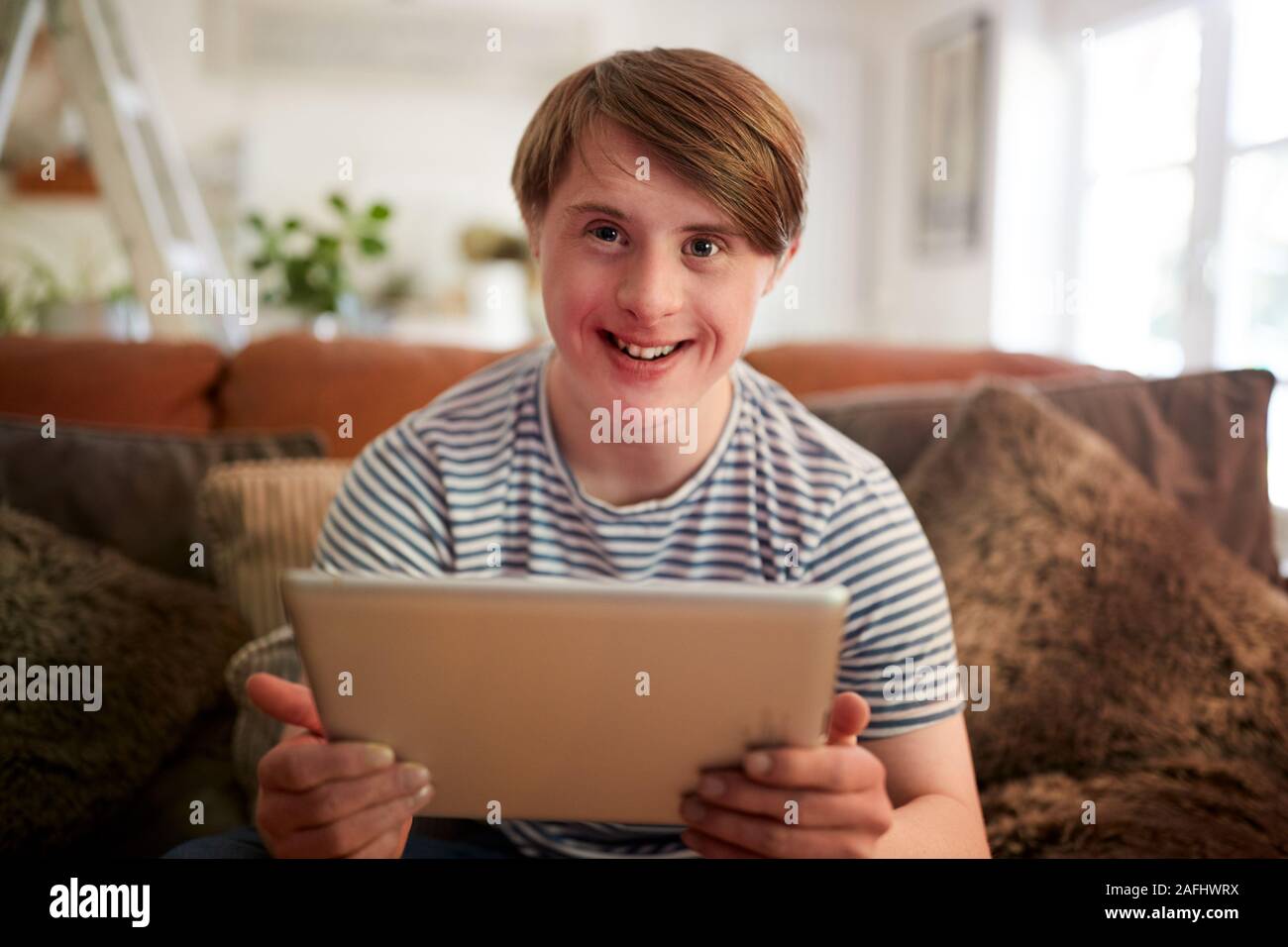 Portrait Of Young Downs Syndrome Man Sitting On Sofa Using Digital Tablet At Home Stock Photo