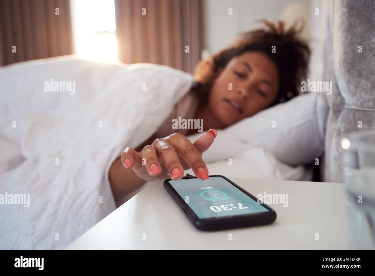 Woman Waking Up In Bed Reaches Out To Turn Off Alarm On Mobile Phone Stock Photo