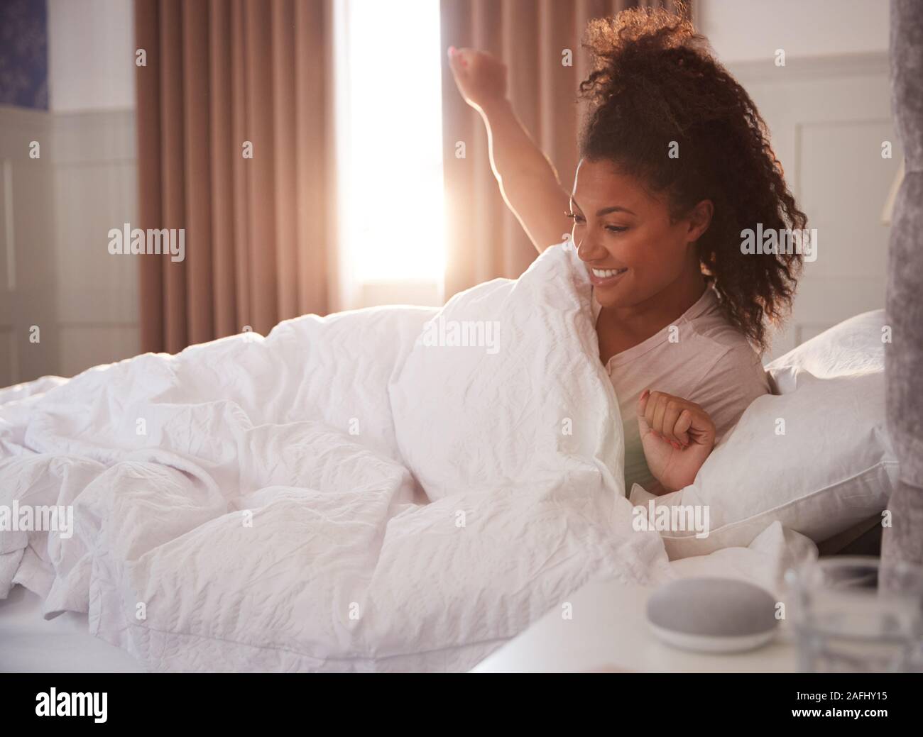 Woman Waking Up In Bed With Voice Assistant On Bedside Table Next To Her Stock Photo