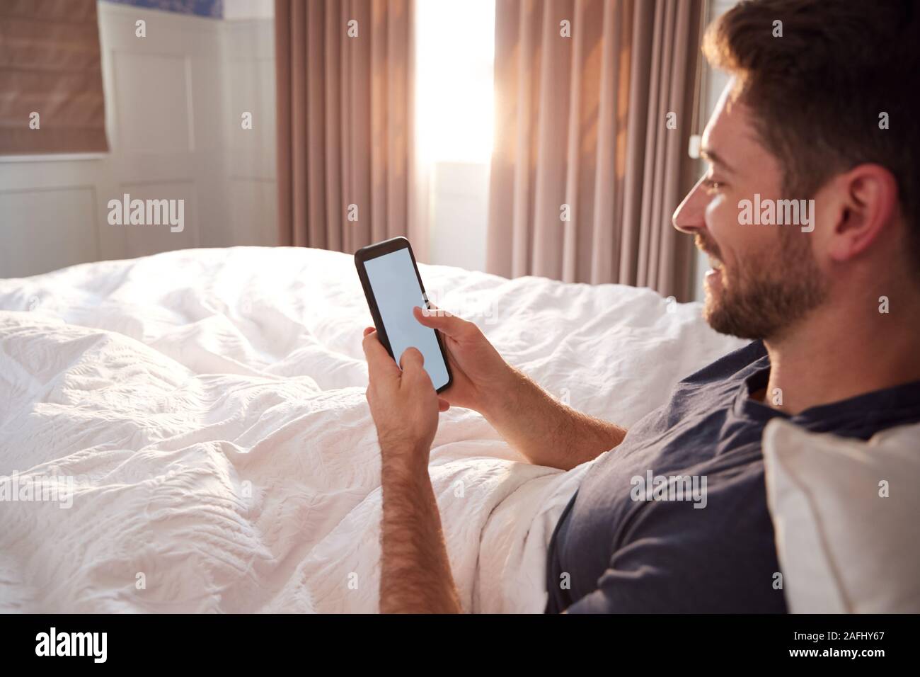 Man Sitting Up In Bed Looking At Mobile Phone After Having Woken Up Stock Photo