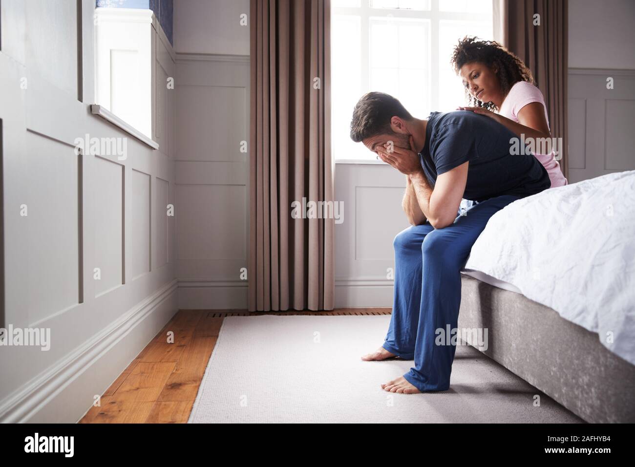 Woman Comforting Man Wearing Pajamas Suffering With Depression Sitting On Bed At Home Stock Photo
