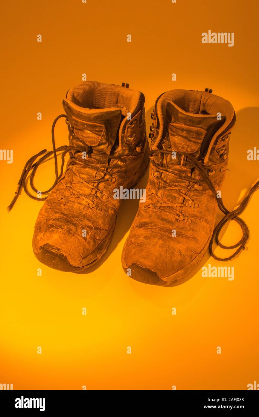 Pair of muddy walking boots bathed in orange pool of light. Metaphor hot footed perhaps, or tired feet, pounding the beat, abstract footwear. Stock Photo