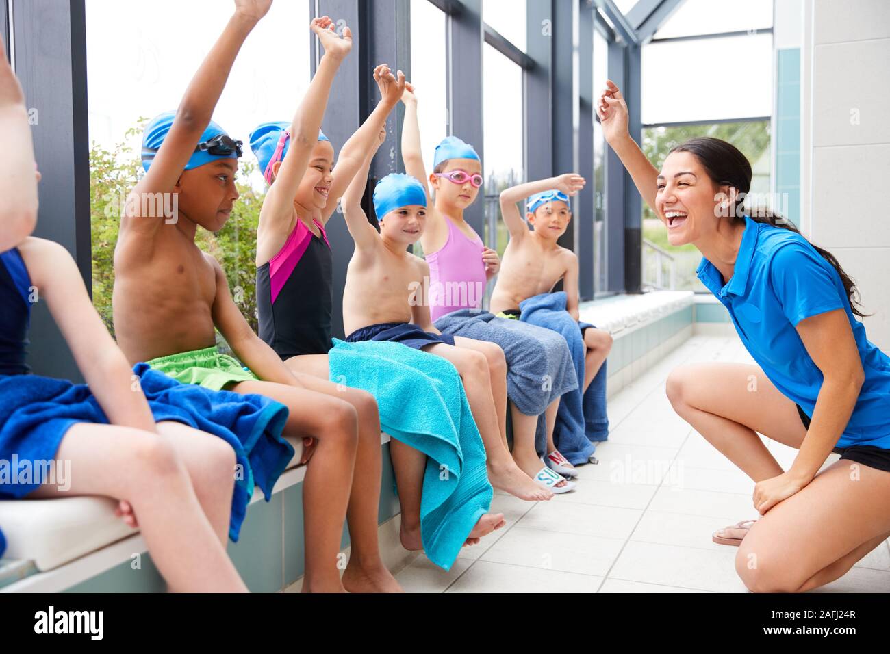 Female Coach Giving Children In Swimming Class Briefing As They Sit On Edge Of Indoor Pool Stock Photo