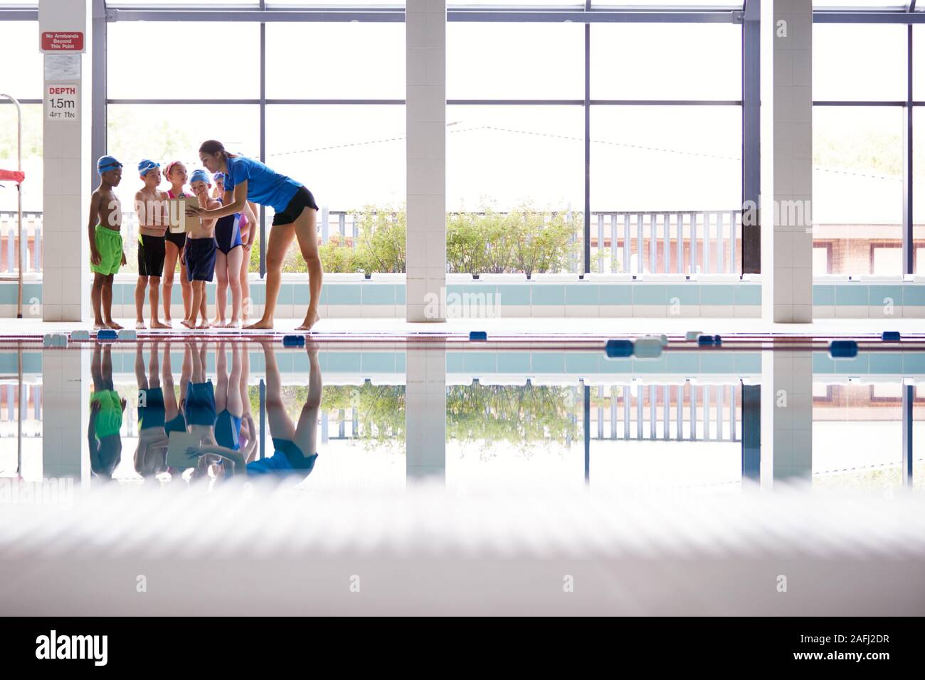 Female Coach Giving Children In Swimming Class Briefing As They Sit On Edge Of Indoor Pool Stock Photo