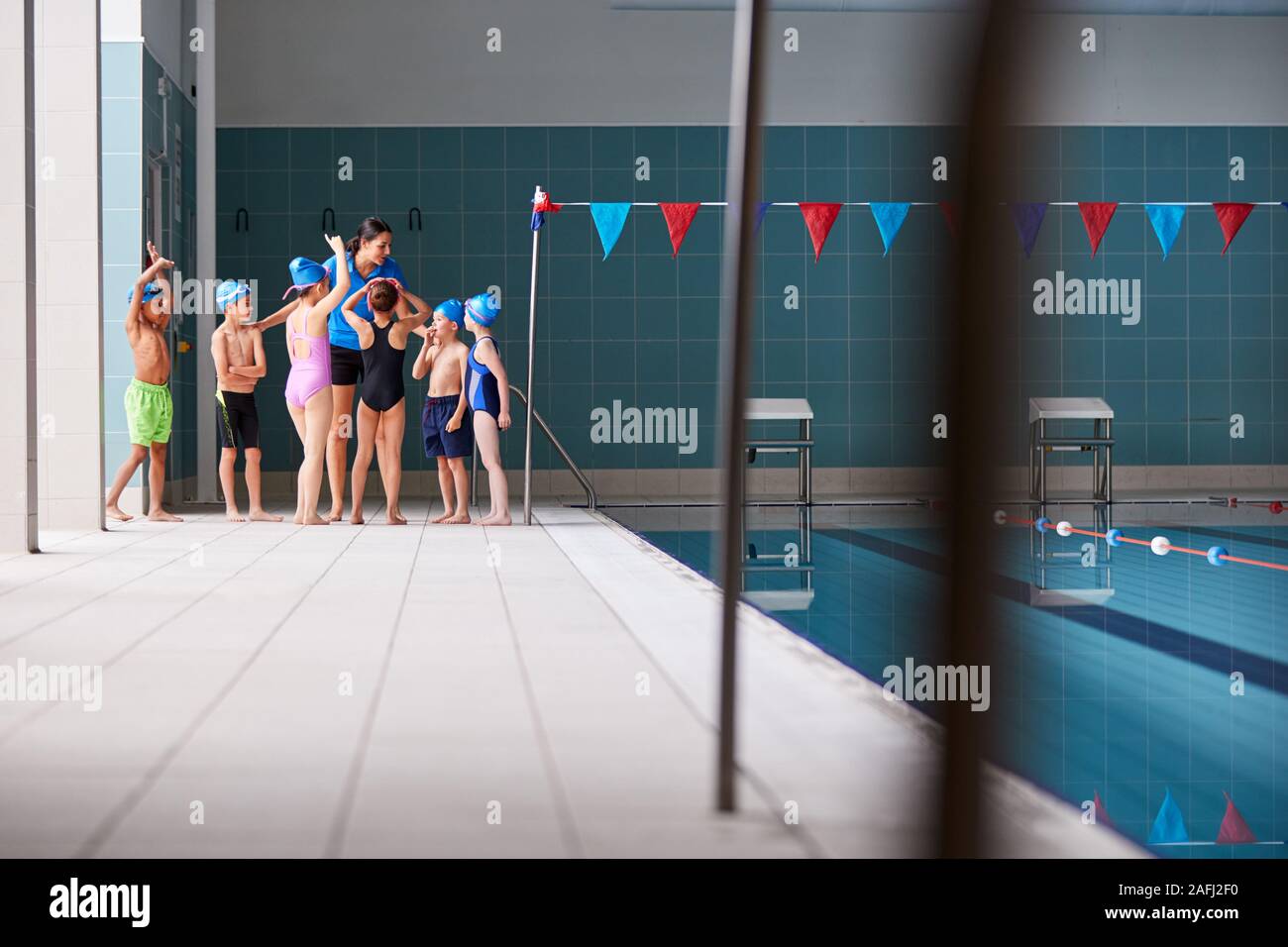 Female Coach Giving Children In Swimming Class Briefing As They Sit On Edge Of Indoor Pool Stock Photo