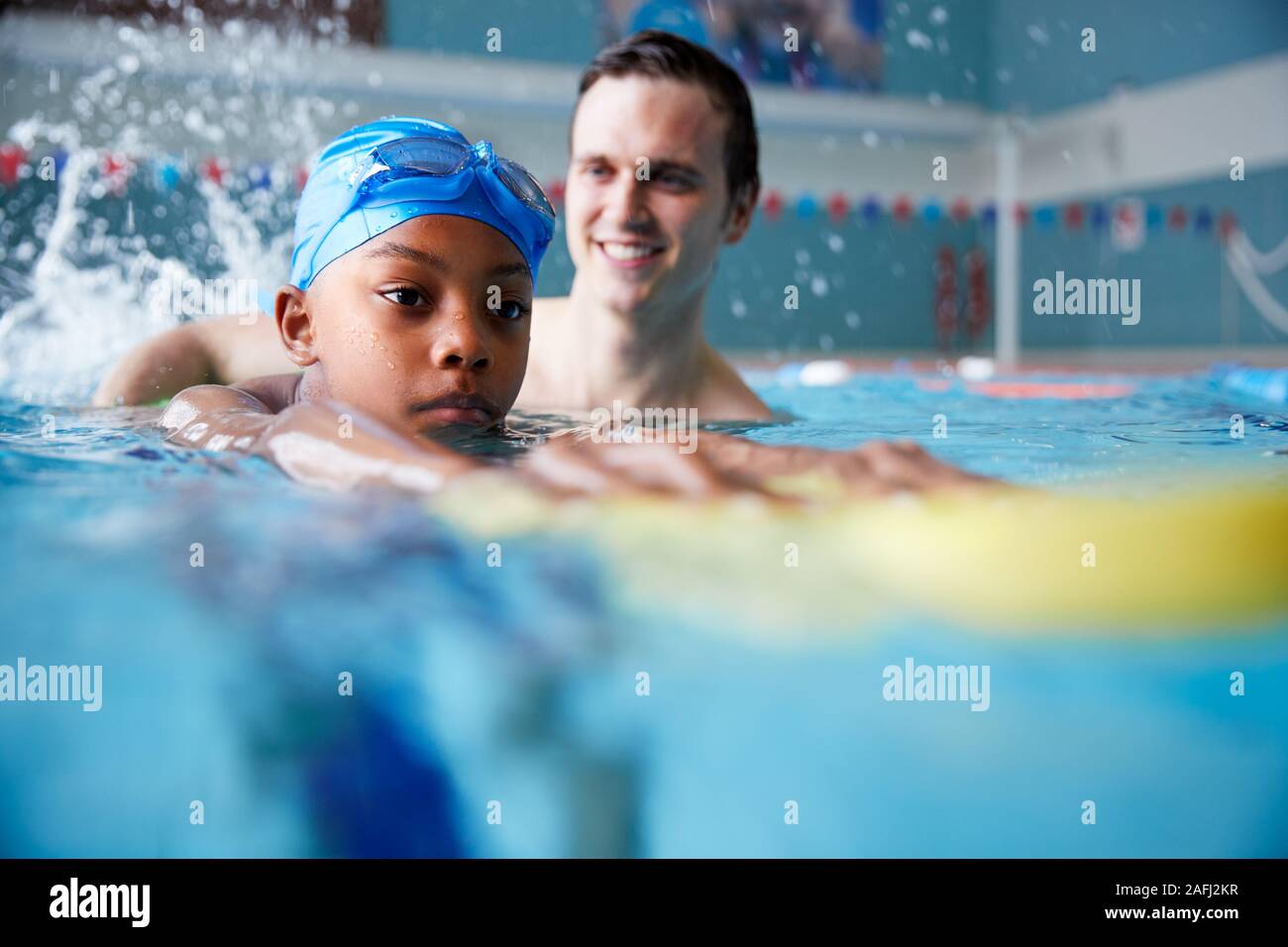 Male Swimming Coach Giving Boy Holding Float One To One Lesson In Pool