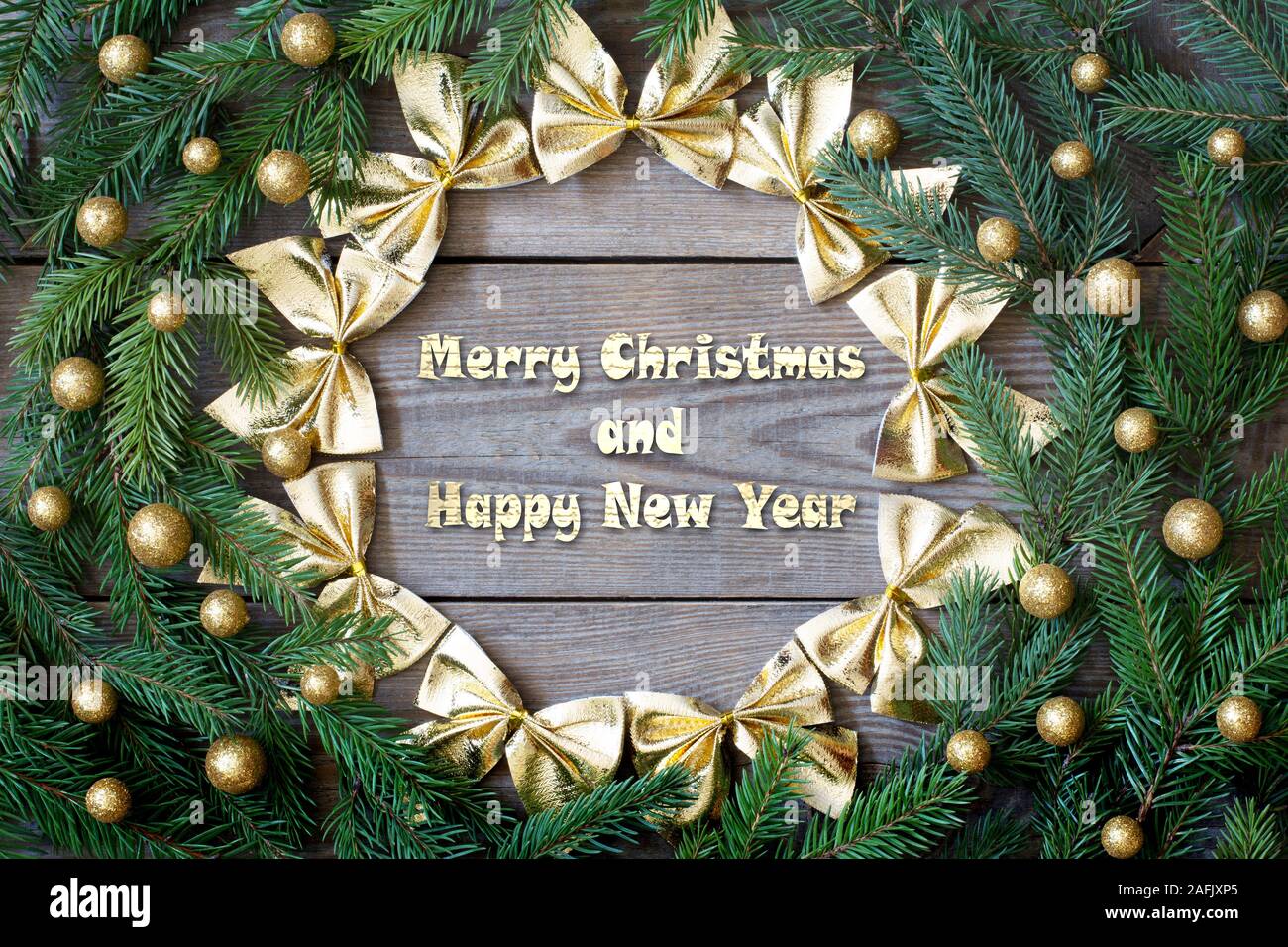 Christmas card concept with fir tree branches, brocade globules and a circle of golden bows on wooden background - greetings text Stock Photo