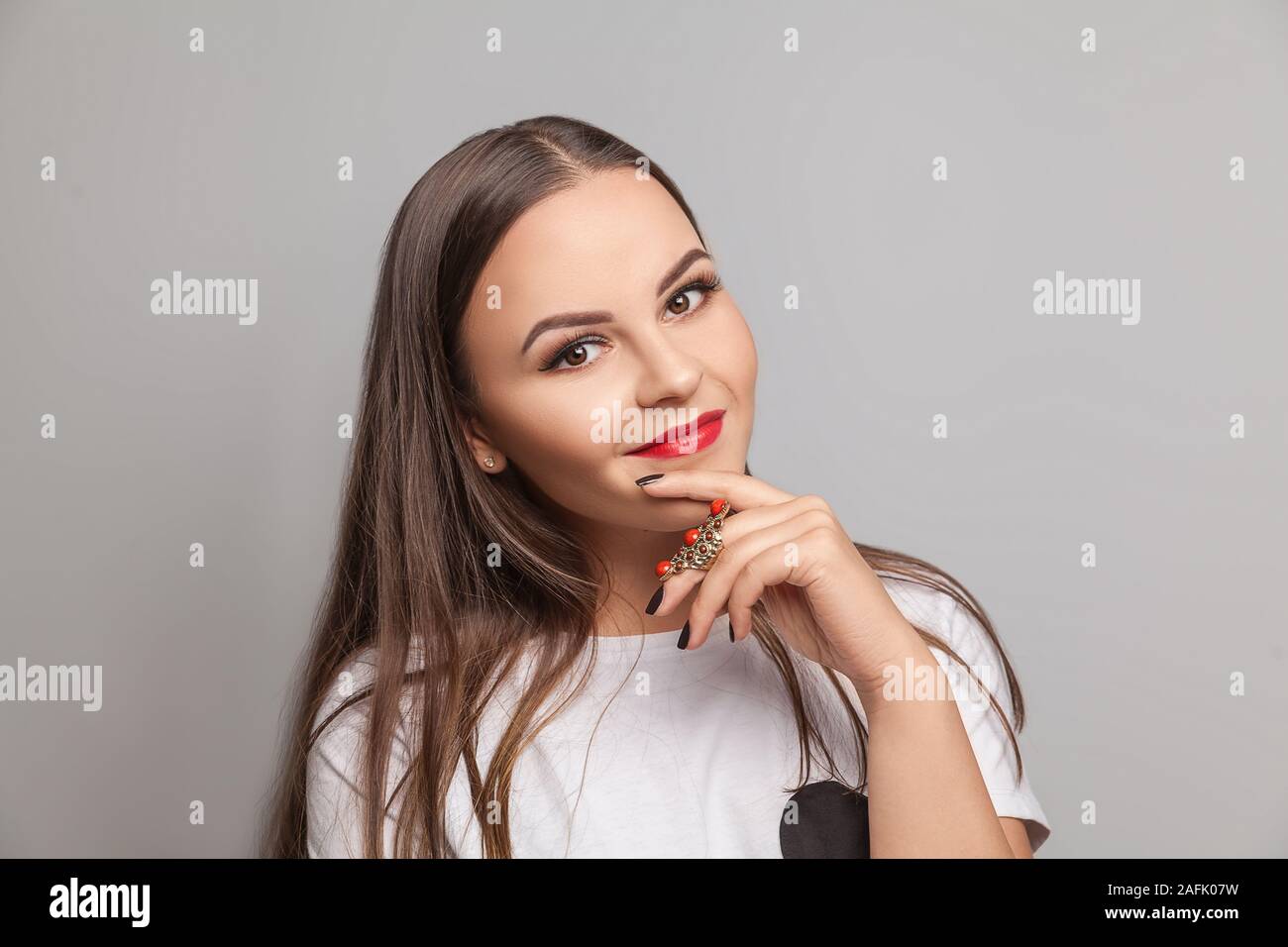 Portrait of young sexy playful brunette girl dressed in white t-shirt standing against isolated gray background. Woman looking at camera. Stock Photo