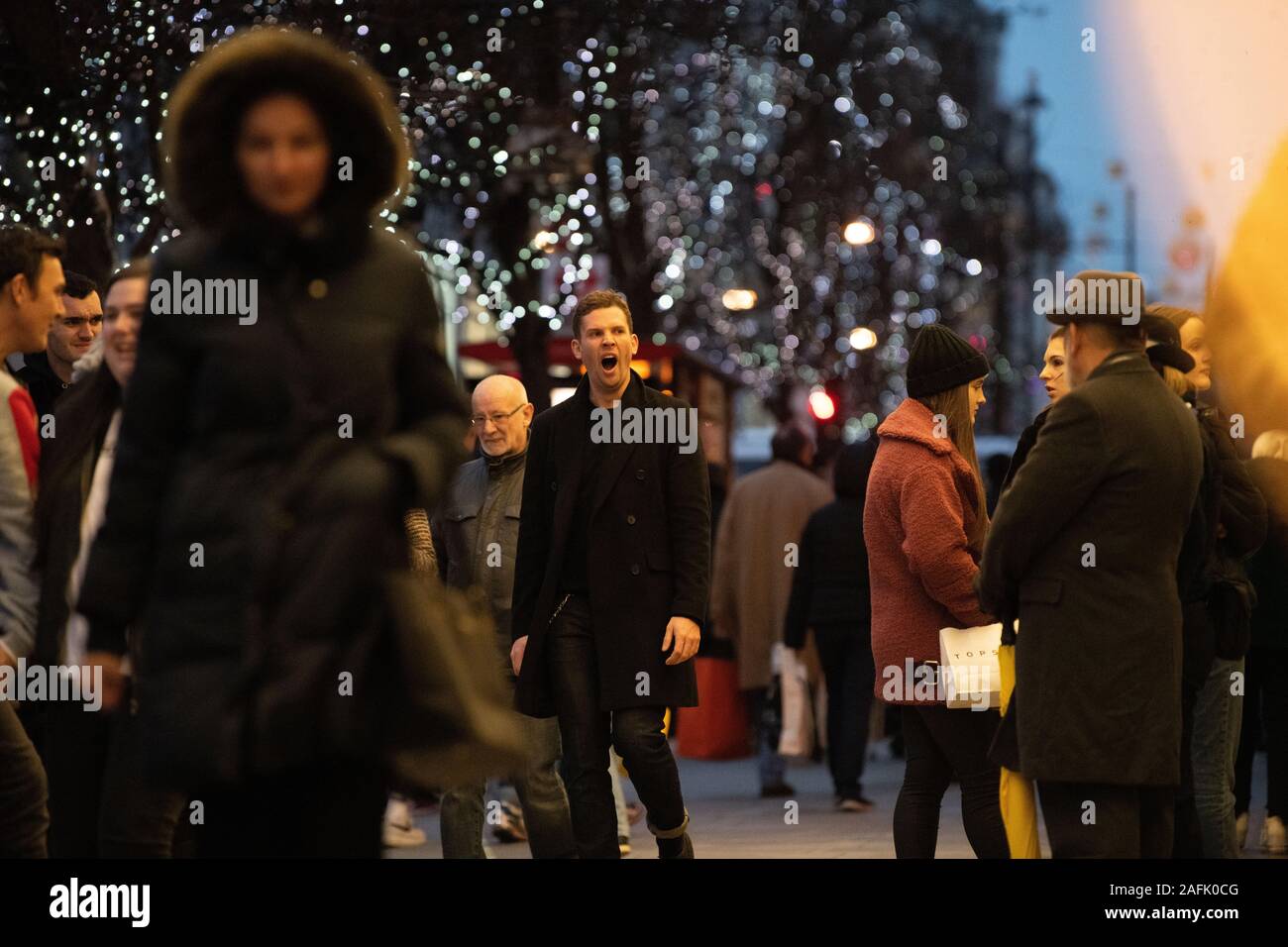 Man in a crowd of Christmas shoppers walking and yawning outside Selfridges, Oxford Street, London, UK. Christmas lights in view Stock Photo