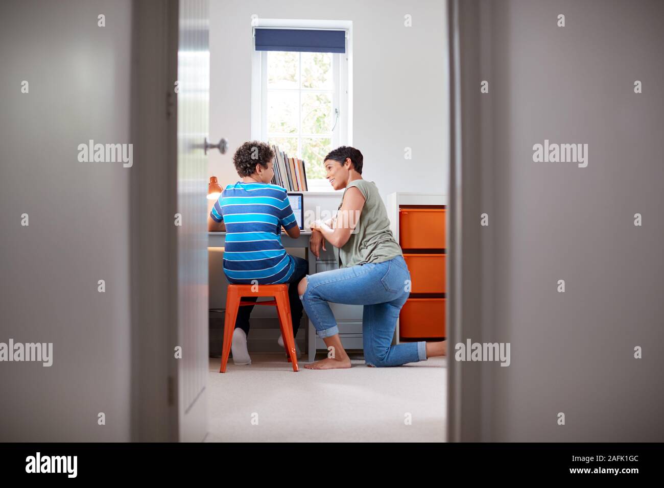 View Through Bedroom Door As Mother Helps Son Using Laptop On Desk To