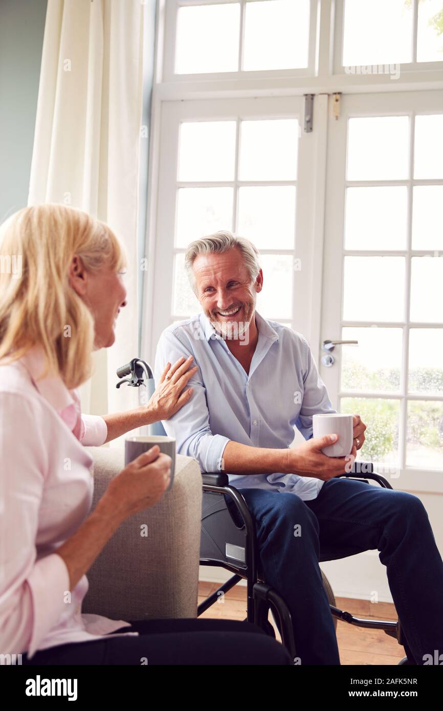 Mature Couple With Man In Wheelchair Sitting In Lounge At Home Talking Together Stock Photo