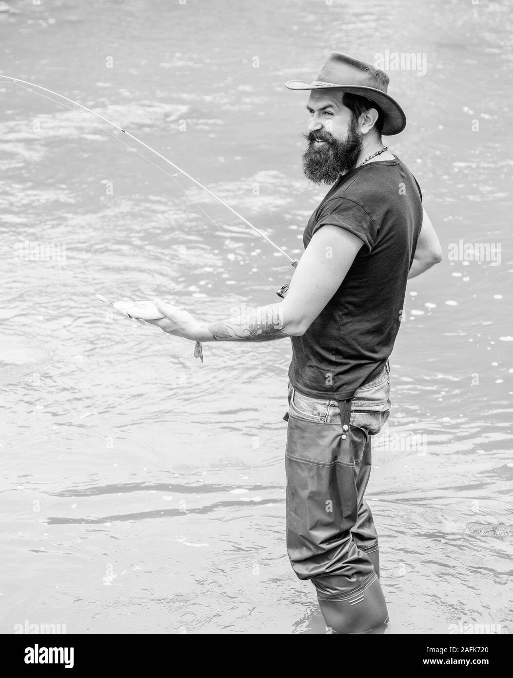 Fun of fishing is catching. Fisher with fishing equipment. Fish on hook. Leisure in wild nature. Fishing masculine hobby. Brutal man wear rubber boots stand in river water. Fisher weekend activity. Stock Photo