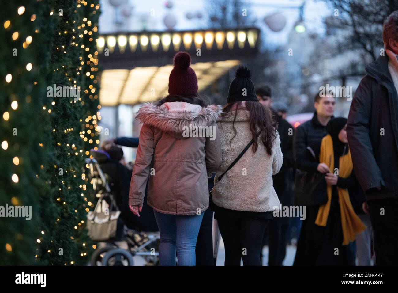 Back of two people wearing winter hats with a bobble walking  towards the iconic entrance of Selfridges, Oxford Street, London, Winter lights in view. Stock Photo