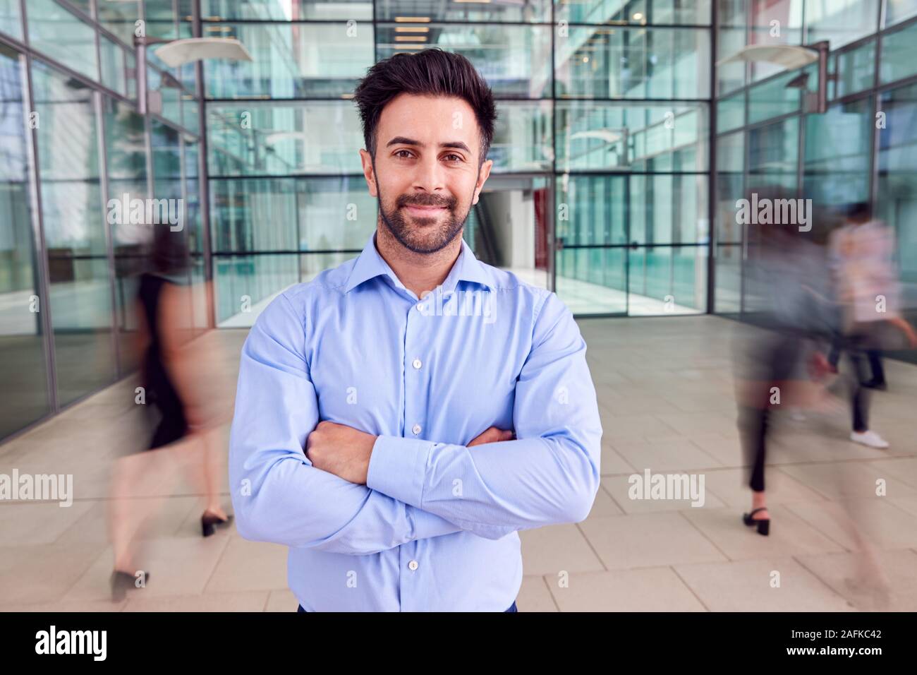 Portrait Of Businessman With Crossed Arms Standing In Lobby Of Busy ...
