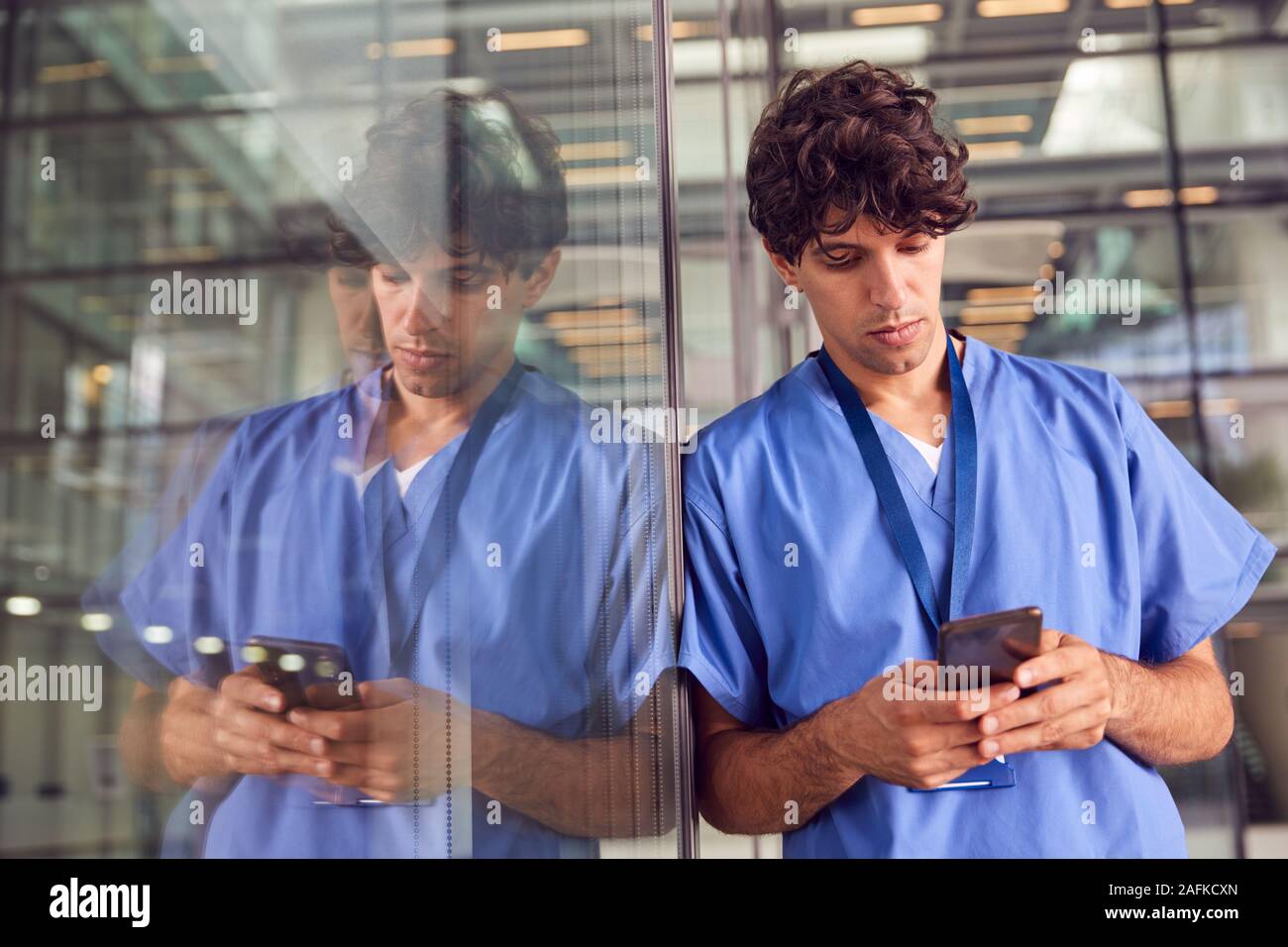 Young Male Doctor Wearing Scrubs Using Mobile Phone Leaning Against Wall In Modern Hospital Building Stock Photo