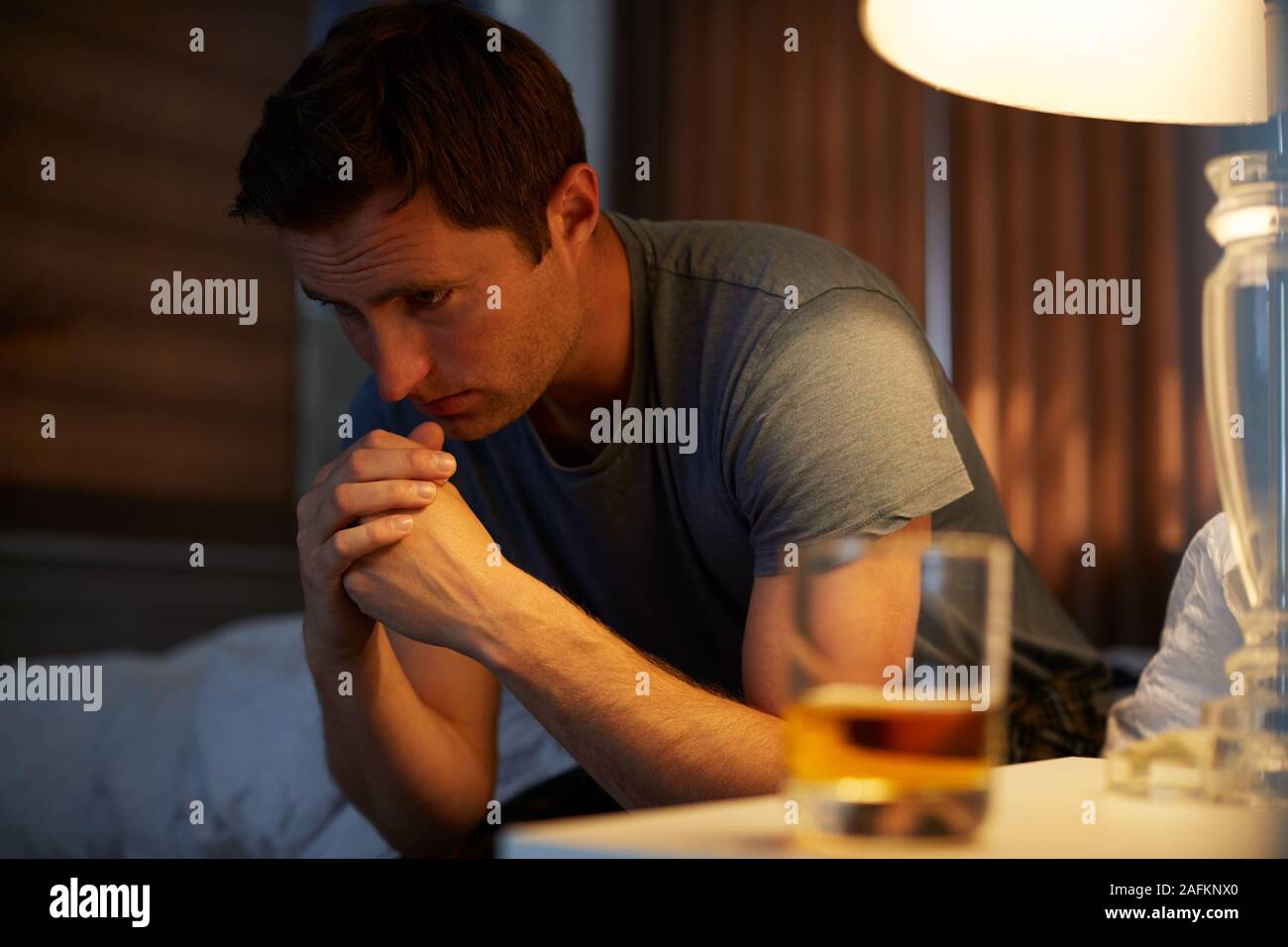 Depressed Man Wearing Pajamas Sitting On Side Of Bed With Glass Of Whisky On Bedside Cabinet Stock Photo