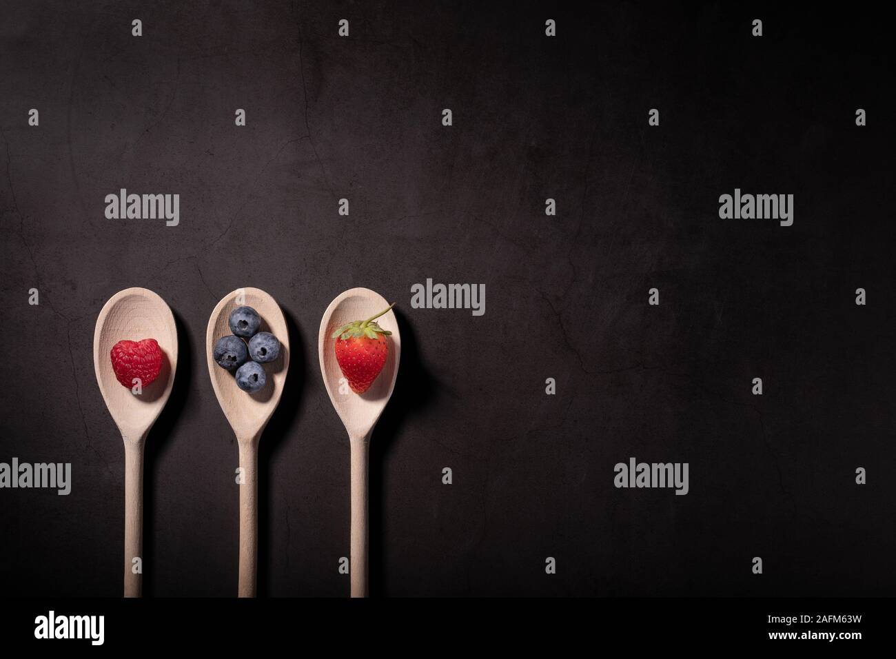 Raspberry, Blueberries and a Strawberry on wooden spoons photographed on a dark background with copy space. Stock Photo