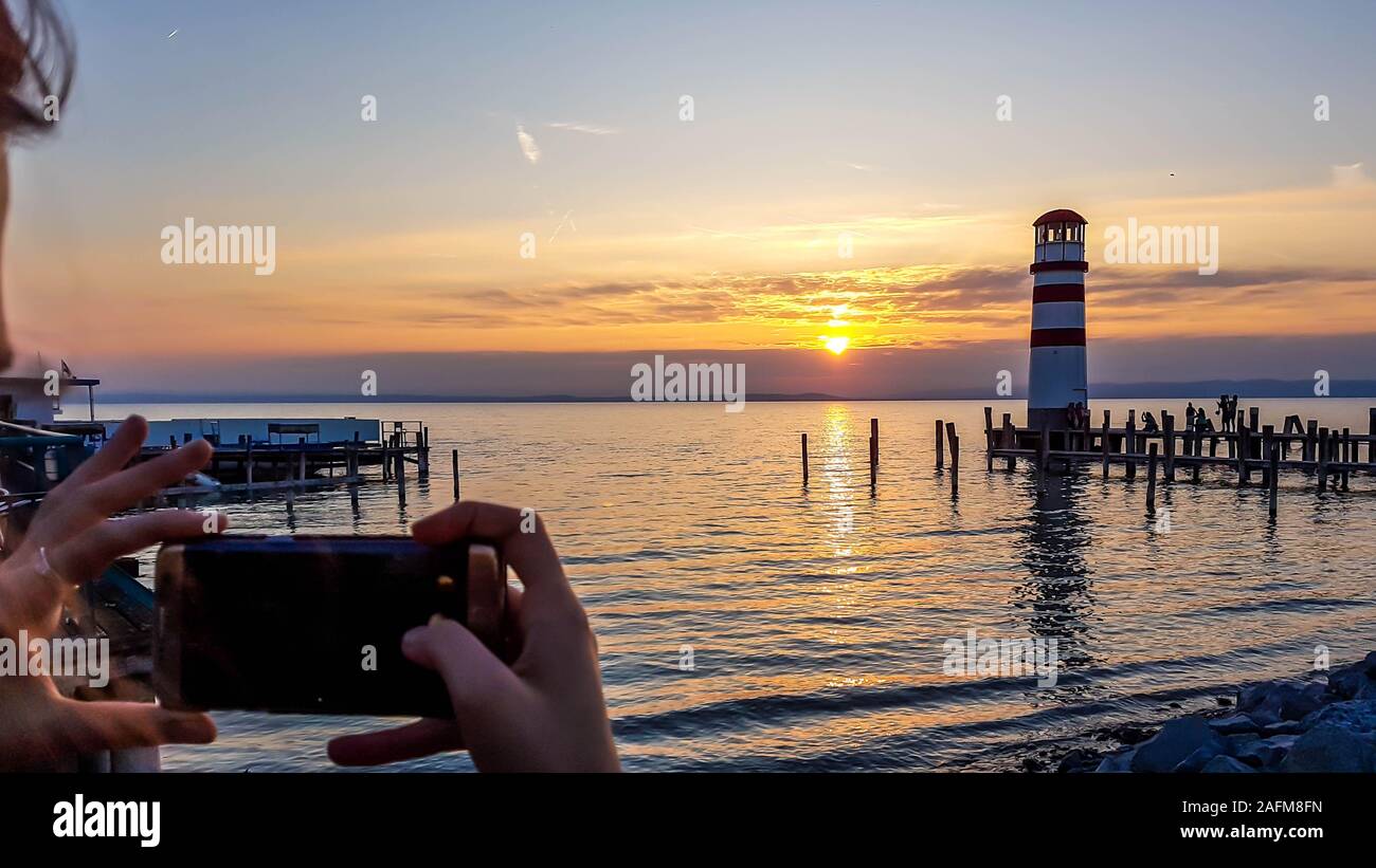A young woman taking picture of a lighthouse located on the lake. Lighthouse has white and red stripes. Sun reflects itself in the calm water of the l Stock Photo