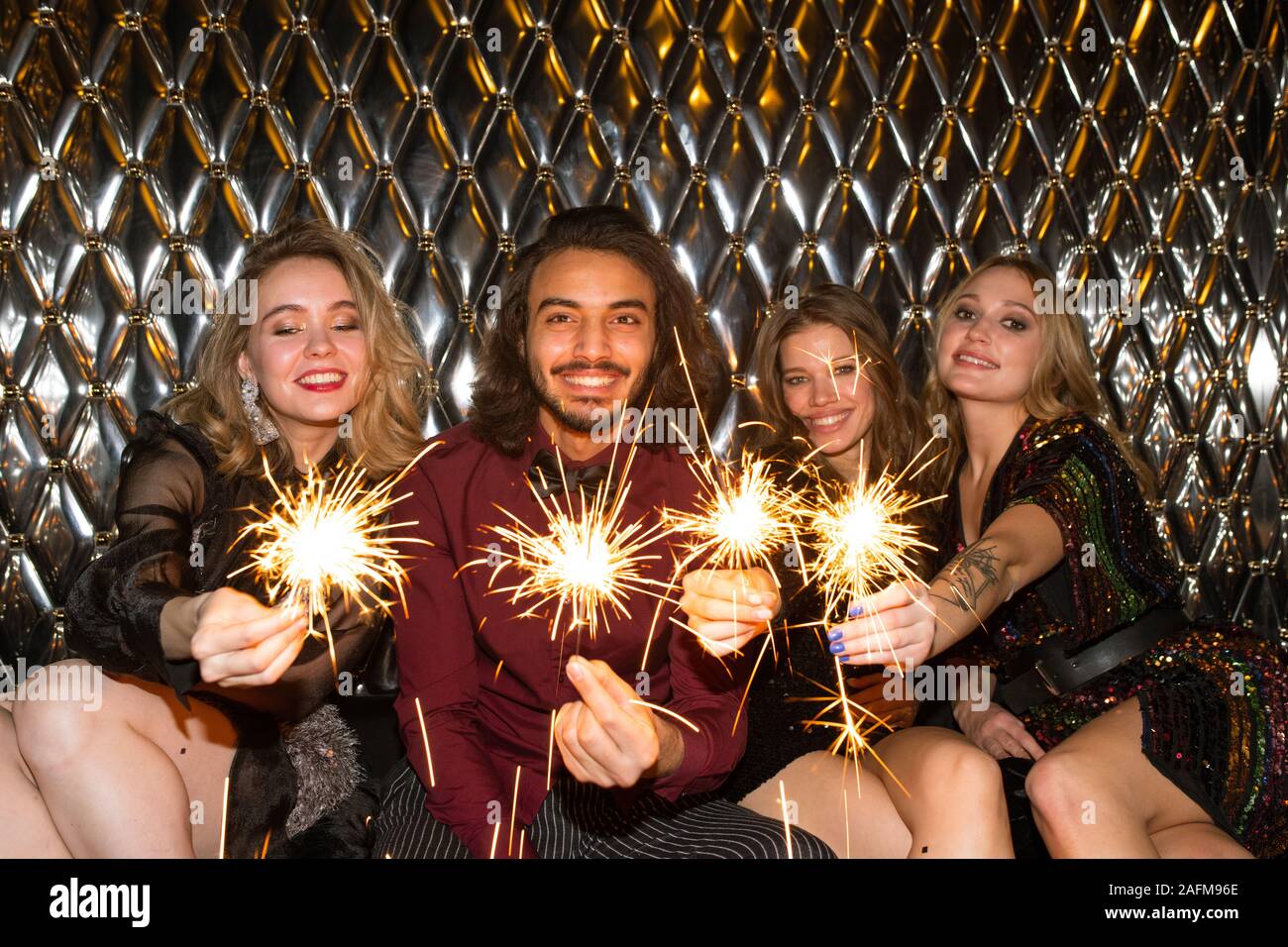 Happy girls and guy with sparkling bengal lights sitting by wall in night club Stock Photo