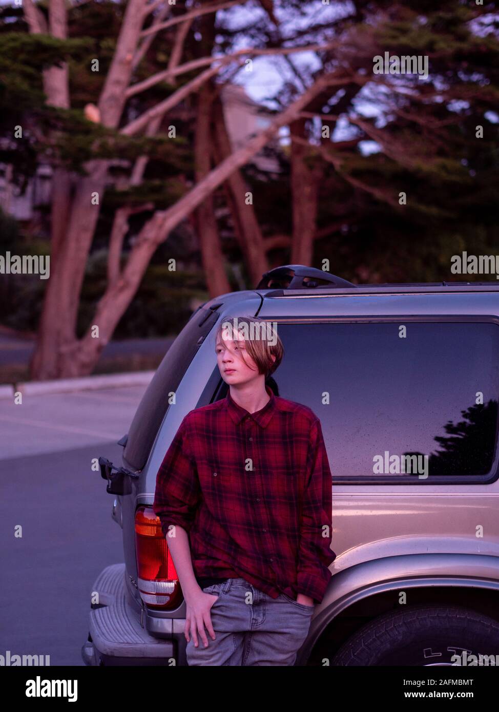 Teenager leaning against SUV looking off frame at sunset Stock Photo