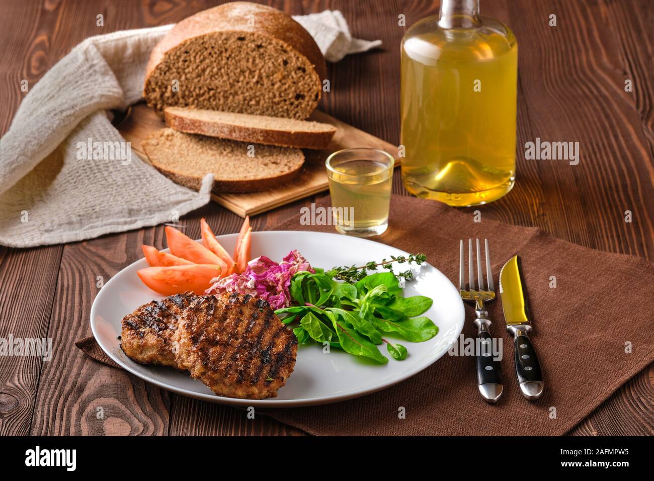 Grilled beef cutlet with fresh salad, brown bread and moonshine on rustic wooden table Stock Photo