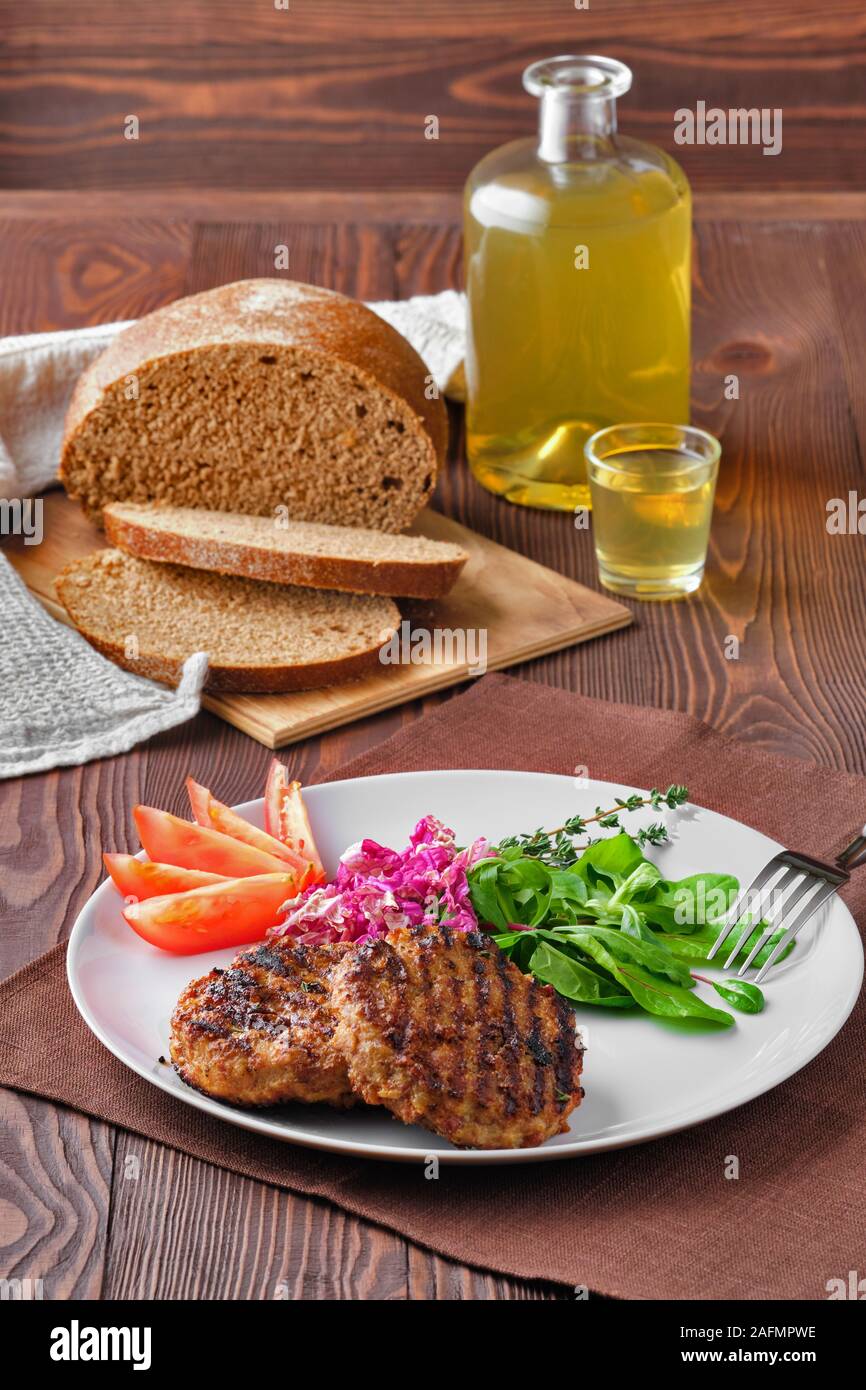Grilled beef cutlet with fresh salad, brown bread and moonshine on rustic wooden table Stock Photo