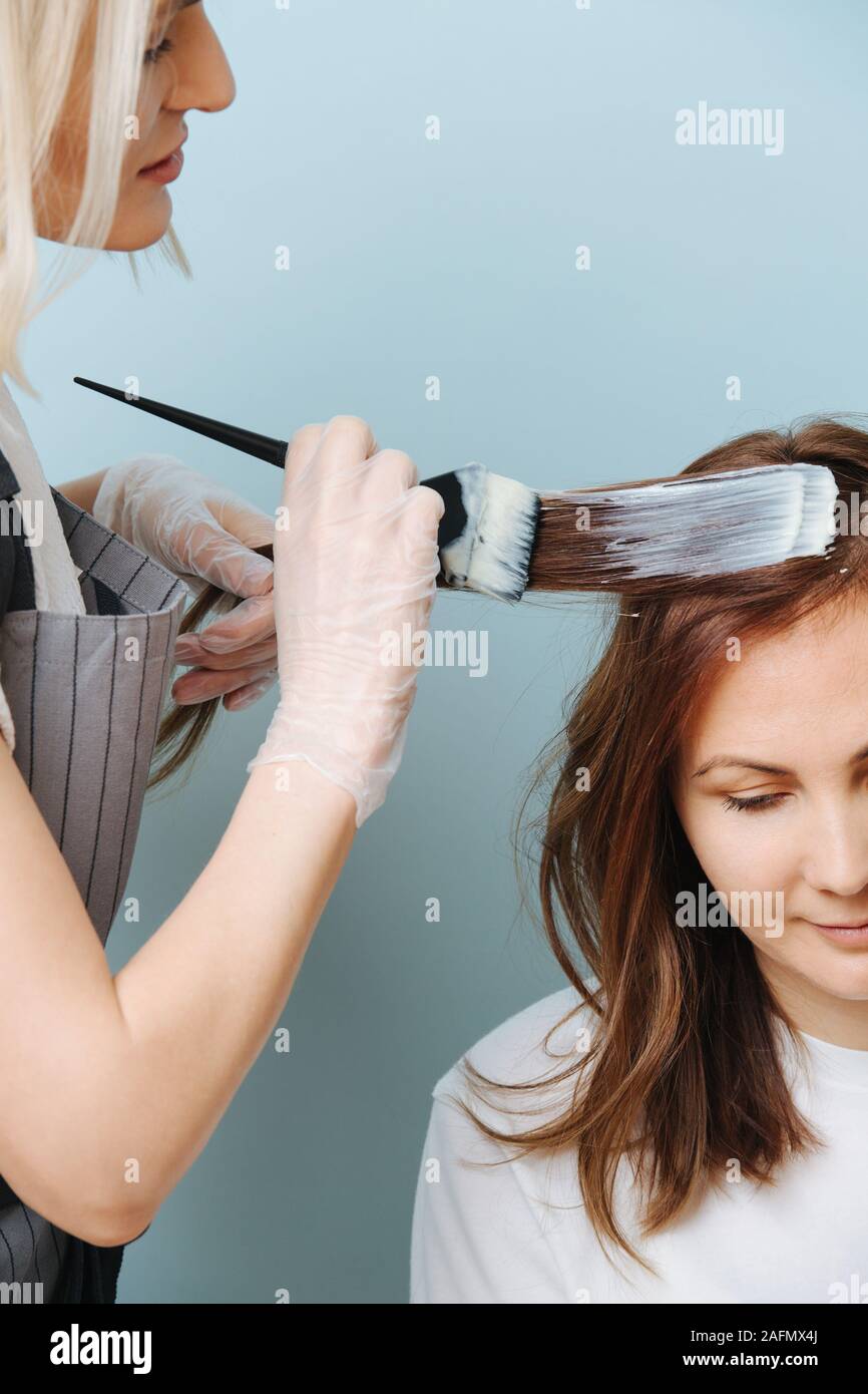 Blonde woman hairdresser applying developer on client's hair over blue. Stock Photo