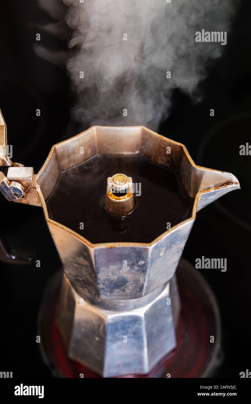 Fresh brewed coffee in a mocha pot on a red hotplate with white steam rising up, close-up, selective focus, vertical format Stock Photo