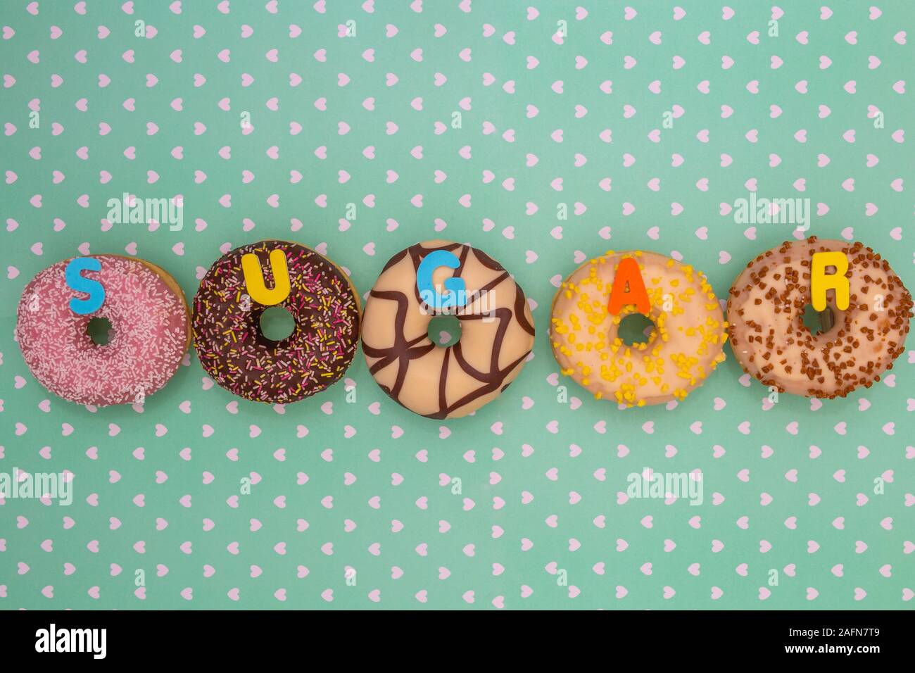 Group of delicious colorful glazed donuts near green background with white hearts pattern and the word sugar Stock Photo