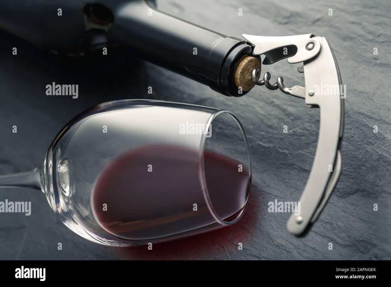 Stainless wine corkscrew in a cork of wine bottle neck and wine glass with wine lying on a black rocky slate background Stock Photo