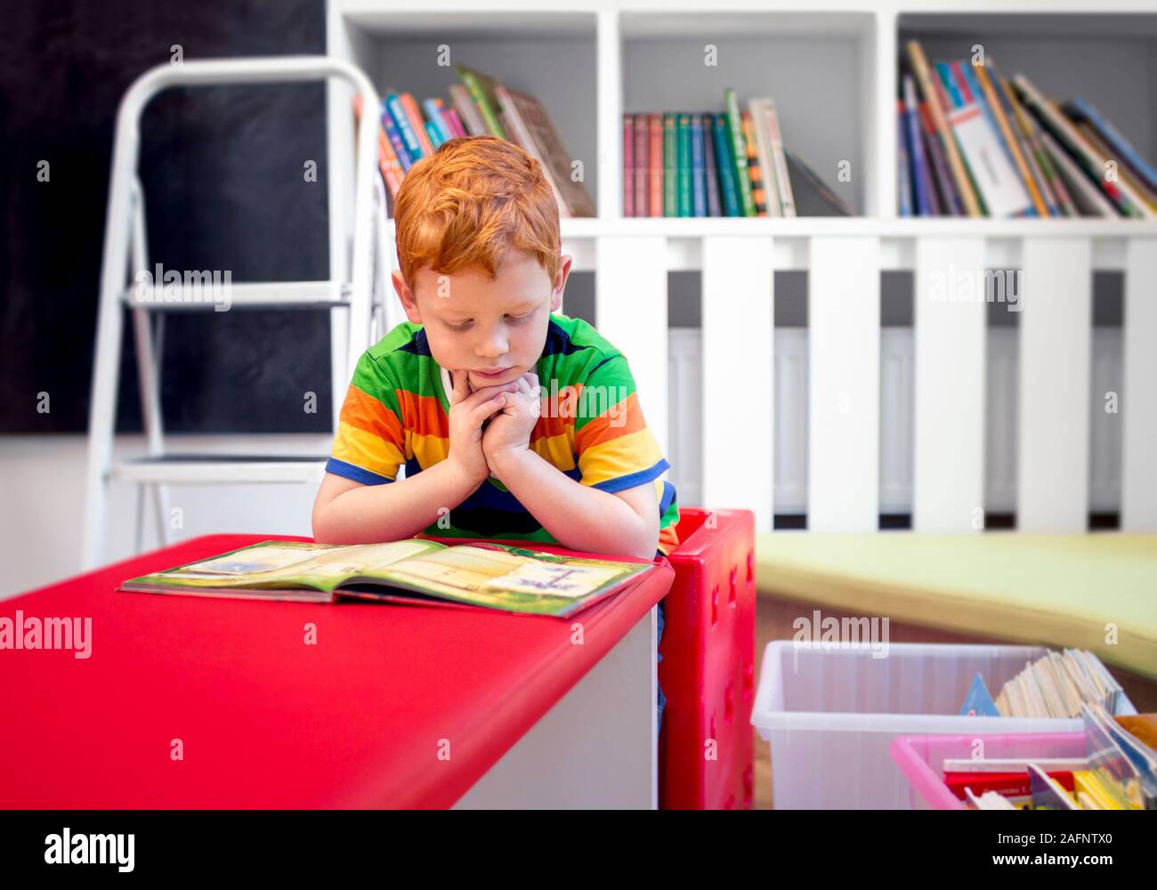 Adorable red-haired little boy, reading book in the library. Bookshelves. Student in preschool reading a magazine book. Education, knowledge. Kid and Stock Photo