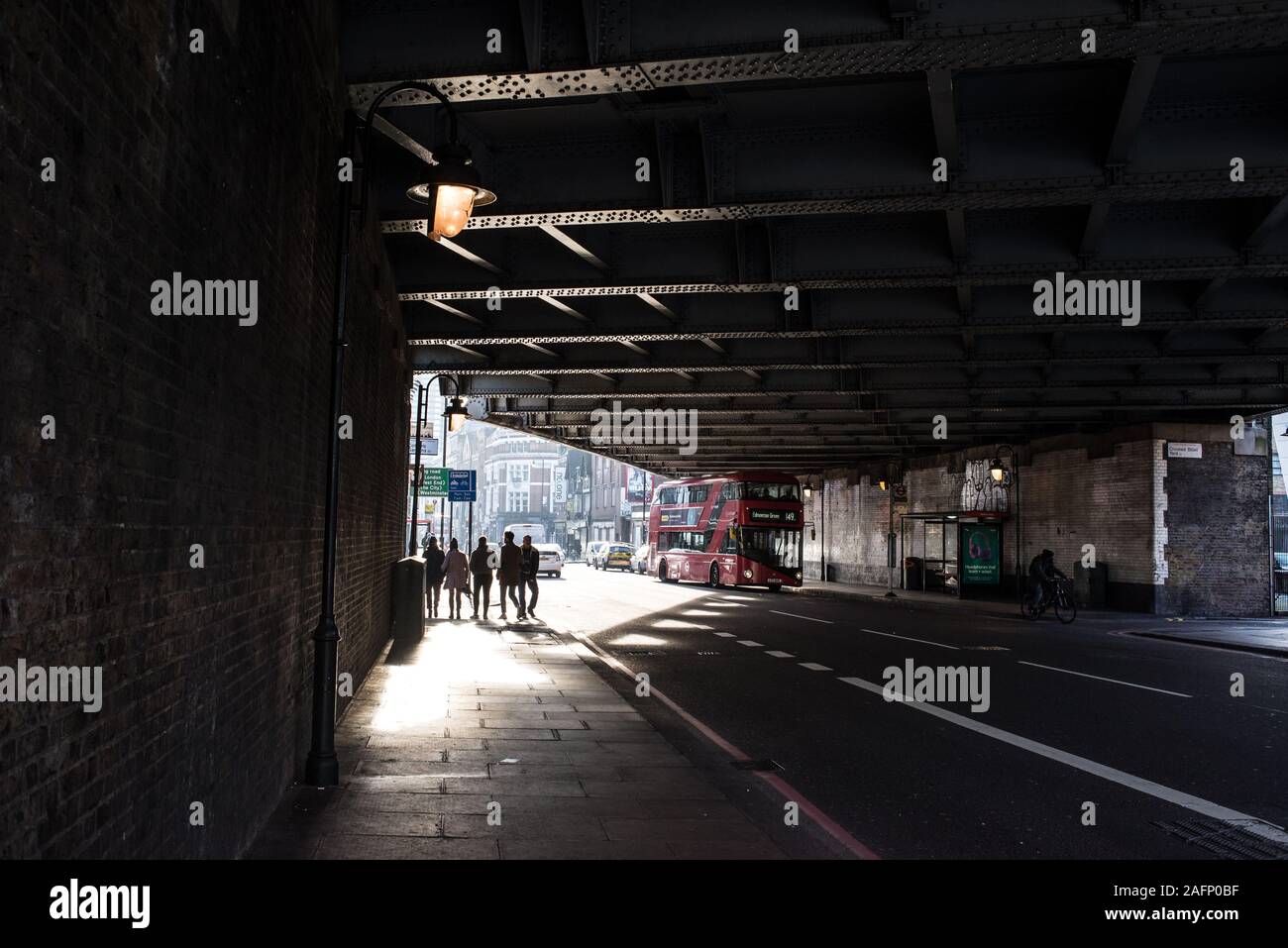 Street scene in Shoreditch, East London with shadows silhouettes of five people walking under a road bridge and red London bus Stock Photo