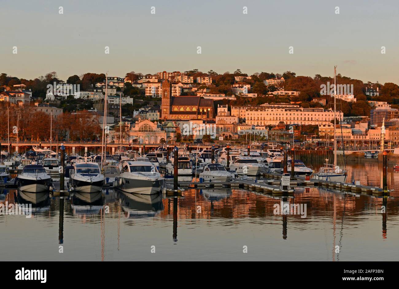 Yachts and other boats moored in the marina at Torquay, Devon, UK, as the sun sets, illuminating the town behind with orange light. Stock Photo